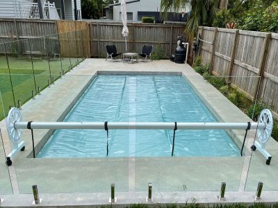 Pool Express - A rectangular backyard swimming pool covered with a pool cover, surrounded by a glass fence, with patio furniture and a barbecue in the background.