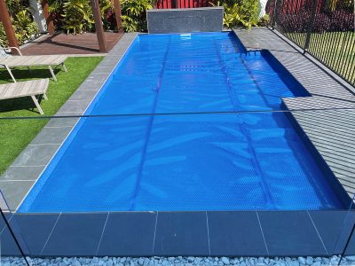Pool Express - Rectangular backyard swimming pool covered with a blue pool cover, surrounded by gray tiles, artificial grass, and a fenced area.