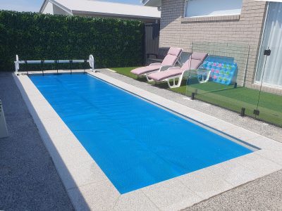 Pool Express - A rectangular outdoor pool with a blue cover, two lounge chairs with cushions, and a glass fence on artificial grass beside a brick house.