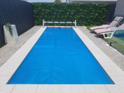 Pool Express - Rectangular swimming pool with a blue pool cover, surrounded by gravel and stone tiles, with two lounge chairs and a decorative plant nearby.