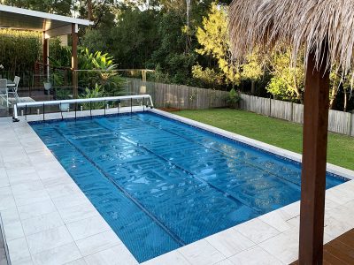Pool Express - A backyard swimming pool with a blue cover partially rolled up, surrounded by a tiled patio, lawn, plants, and a wooden fence.