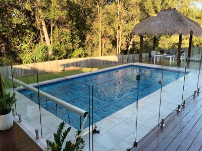 Pool Express - A rectangular swimming pool with a glass fence, surrounded by trees, a thatched-roof gazebo, and outdoor furniture on a tiled patio.