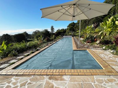 Pool Express - A rectangular backyard pool covered with a blue pool cover, surrounded by stone tiles, plants, and a large white patio umbrella.