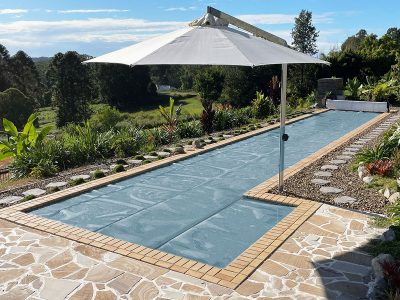 Pool Express - A rectangular backyard pool with a solar cover, bordered by stone and tile, with a large white umbrella providing shade. Lush greenery and trees are visible in the background.
