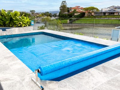 Pool Express - Outdoor pool with a partially unrolled blue pool cover, surrounded by gray stone tiles, next to a waterway and green lawns with houses in the background.