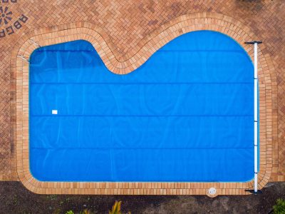 Pool Express - Aerial view of a swimming pool with a blue cover, surrounded by a brick patio. The pool has a curved rectangular shape.