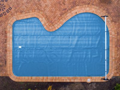 Pool Express - Aerial view of a kidney-shaped swimming pool covered with a blue pool cover, surrounded by a brick-paved deck.