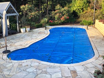 Pool Express - Rectangular swimming pool outdoors with a blue safety cover, surrounded by stone paving, trees, and shrubs in the background.