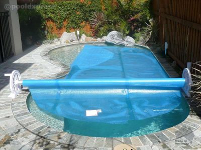 Pool Express - A residential backyard pool partially covered with a blue bubble pool cover on a rolling reel, surrounded by stone tile and garden plants.