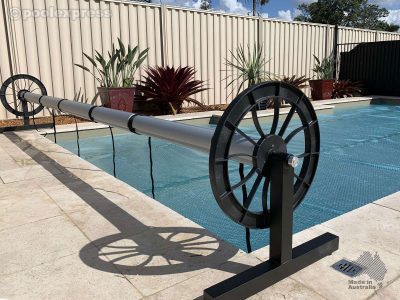 Pool Express - A pool cover roller with a silver tube and black wheels stands beside a swimming pool on a tiled patio, near potted plants and a beige fence.