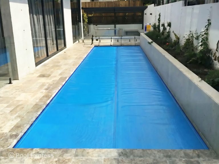 Pool Express - A rectangular swimming pool with a blue pool cover is surrounded by a stone-tiled patio, glass fencing, and a raised garden bed along one side.