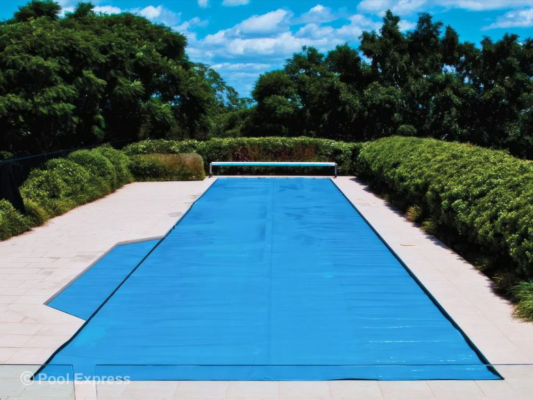 Pool Express - A rectangular outdoor swimming pool is covered with a blue pool cover. The pool is surrounded by a paved area, green hedges, and trees under a sunny sky.