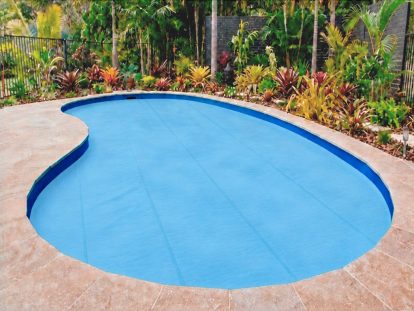 Pool Express - A backyard kidney-shaped pool covered with the 5mm Foam Thermal Cover, surrounded by tiled decking and tropical plants, creates a serene retreat while also ensuring safety—an important consideration when exploring pool cover price options.