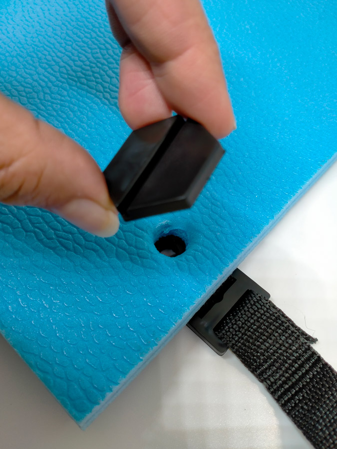 Pool Express - Hand holding a black plastic piece above a hole in blue foam, with a black strap and buckle attached to the foam.