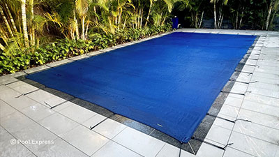 Pool Express - A rectangular pool covered with a blue safety cover, secured with straps, is surrounded by tiled pavement and tropical plants.