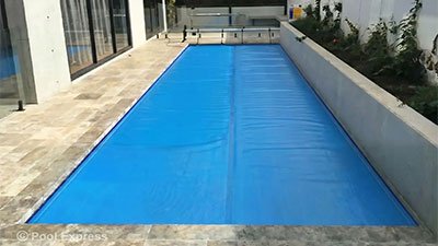 Pool Express - A rectangular swimming pool with a blue pool cover, surrounded by stone tiles and adjacent to a raised garden bed.